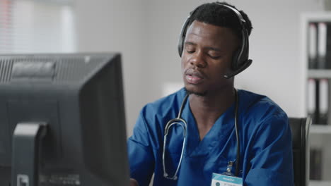 A-black-man-sits-at-a-computer-in-a-doctor's-uniform-and-writes-a-patient's-card-while-taking-calls-with-headphones.-Ambulance-Hotline-receive-calls-and-distribute-ambulances